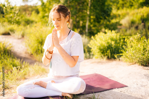 Attractive young woman is meditating and doing breathing exercising sitting in pose of lotus or sukhasna background of beautiful nature, landscape and scenery, holding hand in Namaste gesture.