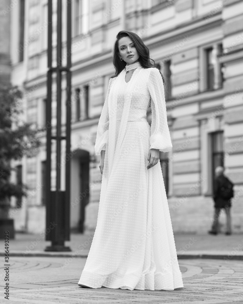 Elegant fashionable stylish female model in long white dress walking at city street. Outdoor full length portrait. Caucasian lady with wavuy hair and makeup