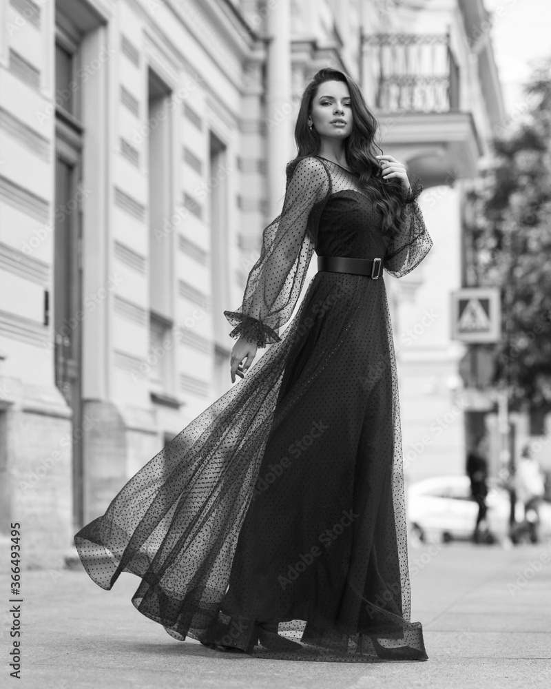 Fototapeta premium Elegant woman wearing long blue evening dress. Pretty lady posing outdoors. Fashionable stylish model. Fashion outdoor portrait. Female with long wavy hair standing at city street