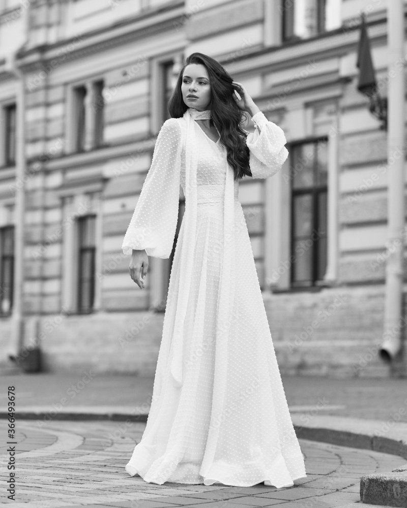 Elegant fashionable stylish female model in long white dress walking at city street. Outdoor full length portrait. Caucasian lady with wavuy hair and makeup