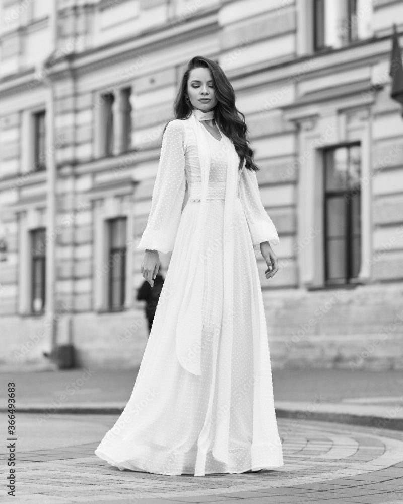 Elegant fashionable stylish female model in long white dress walking at city street. Outdoor full length portrait. Caucasian lady with wavuy hair and makeup Elegant fashionable stylish female model in long white dress walking at city street. Outdoor full length portrait. Caucasian lady with wavuy hair and makeup