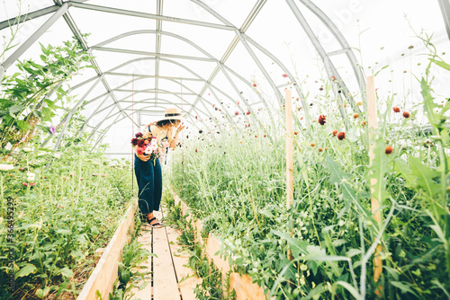 happy young woman in large straw hat with fresh colorful flowers bouquet walks along aromatic blooming plants in modern greenhouse on sunny day