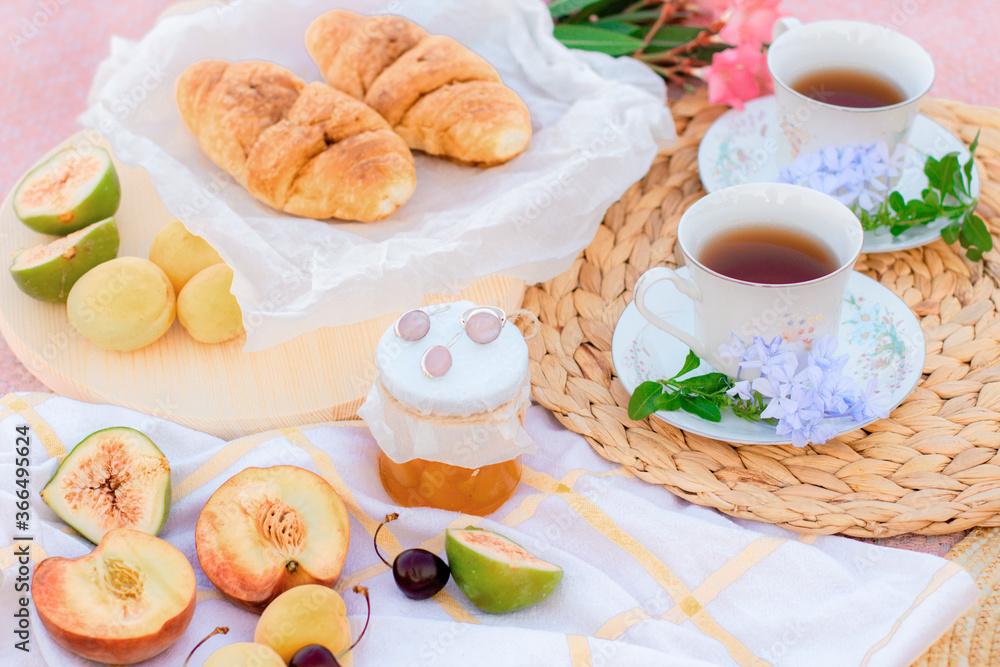 A picnic at dawn in gentle colors. Hot tea, fresh croissants, jam and fruit on a blanket with a wooden stand.