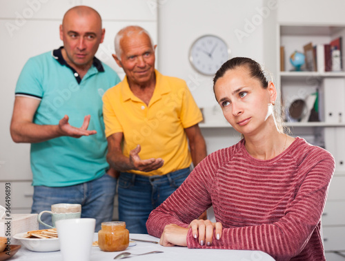 Upset young woman sitting after quarreling with family, men on background at home © JackF