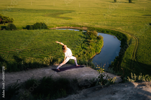 Concentrated young Caucasian woman is performing warrior yoga asana pose on top of rock background of flowing river and green meadow on summer sunny day. Girl practicing Virabhadrasana.