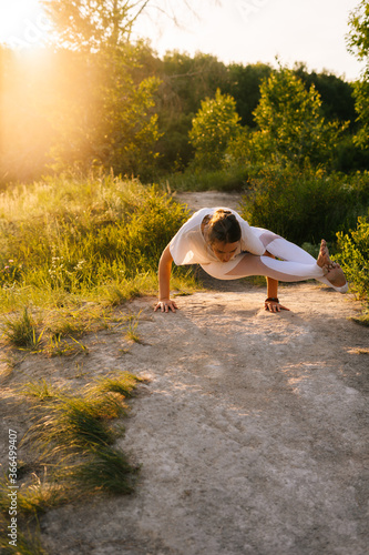 Young woman with stretching body practicing yoga and performing Eight-Angle Pose outside in park evening on background of sunlight. Female doing advanced yoga exercises outdoors at green grass. photo