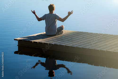 young man on wooden pontoon or pier practicing yoga & relaxing with reflection in water .on holiday