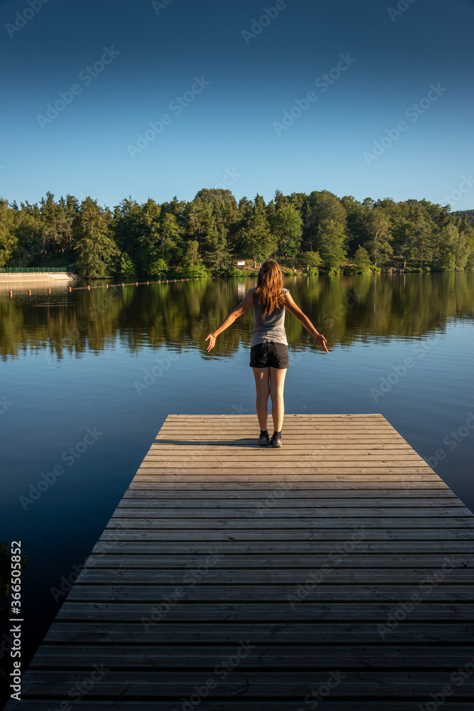 Young woman with outstretched arms standing relaxing or stretching on wooden pontoon staring at lake. evening light Back view .