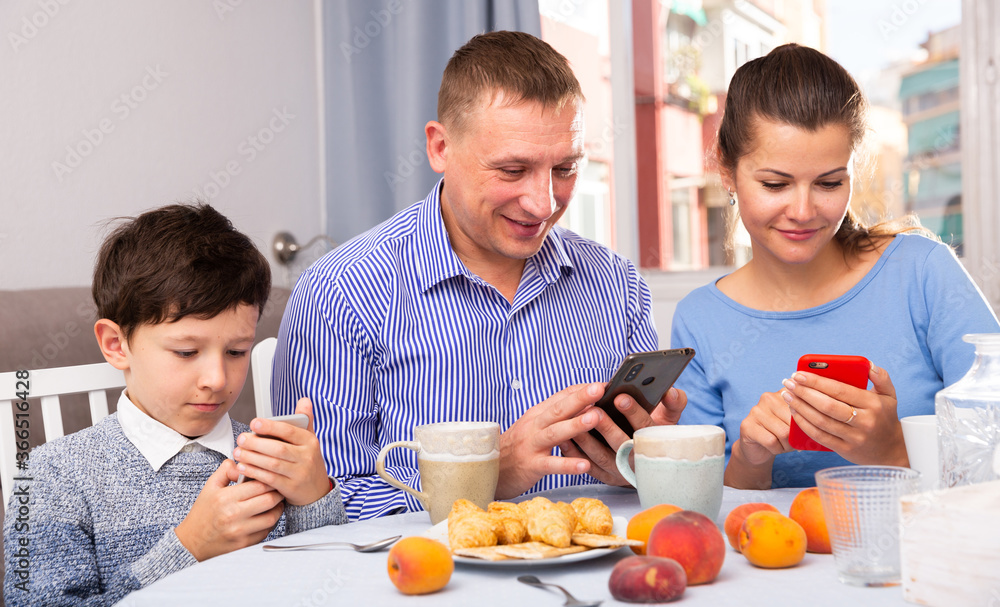 Portrait of cheerful preteen boy and his parents using phones at home..