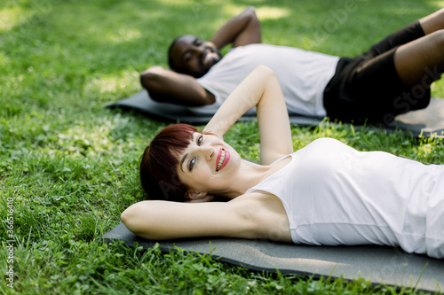 Young fitness multi racial couple in sportswear, doing sit ups on mats in the summer morning at city park, looking at camera with smile. Sport girl doing push up exercise, African man on background