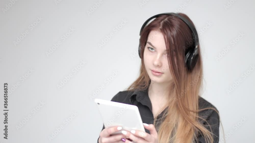 Good looking pretty young girl with long hair in headphones uses small white tablet, white background Good looking pretty young girl with long hair in headphones uses small white tablet, white background