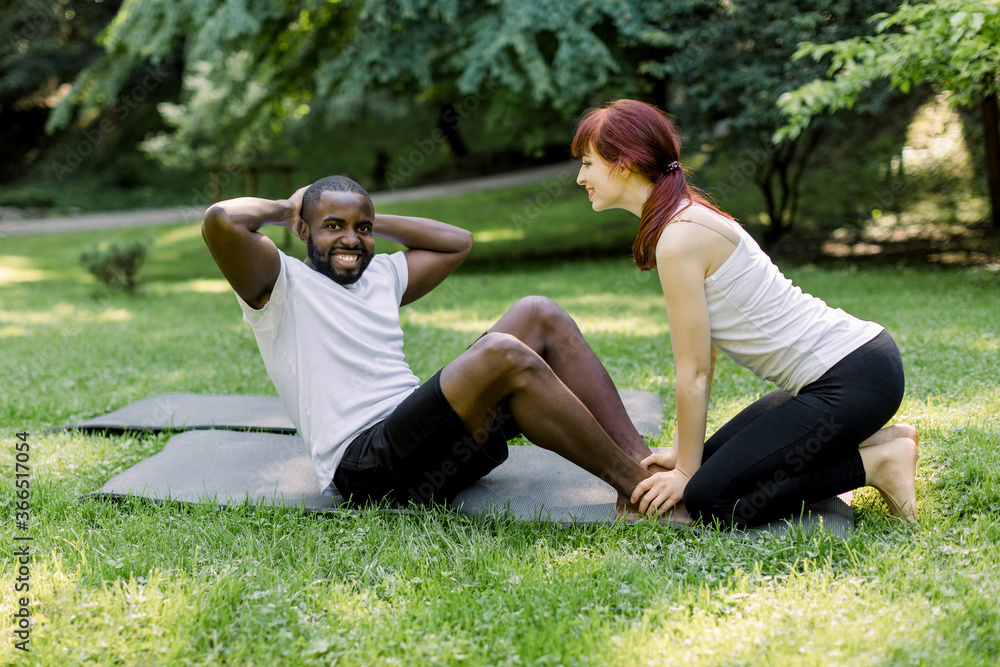 Personal trainer, pretty smiling Caucasian girl, showing abdomen crunches exercise to her male client, joyful African guy. Multiethnical couple doing yoga and fitness exercises in nature at city park. Personal trainer, pretty smiling Caucasian girl, showing abdomen crunches exercise to her male client, joyful African guy. Multiethnical couple doing yoga and fitness exercises in nature at city park.