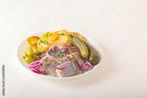Slices of pickled herring with red onion, fermented cucumber, fried potatoes, olives and herbs on a light wooden table. Copy space