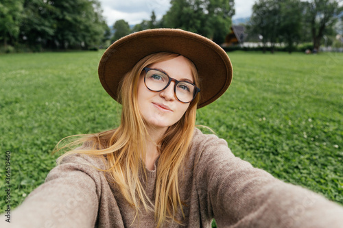 Beautiful young hipster woman taking selfie on green grass at the park. Looking at camera and smile. Travel and active life concept. Outdoors