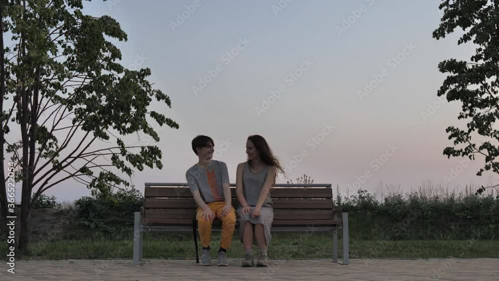 A general shot of a young guy and a modest girl sitting on a bench, looking at each other and laughing merrily, at the end the guy hugs the girl tightly. A couple is sitting in an open space against A general shot of a young guy and a modest girl sitting on a bench, looking at each other and laughing merrily, at the end the guy hugs the girl tightly. A couple is sitting in an open space against