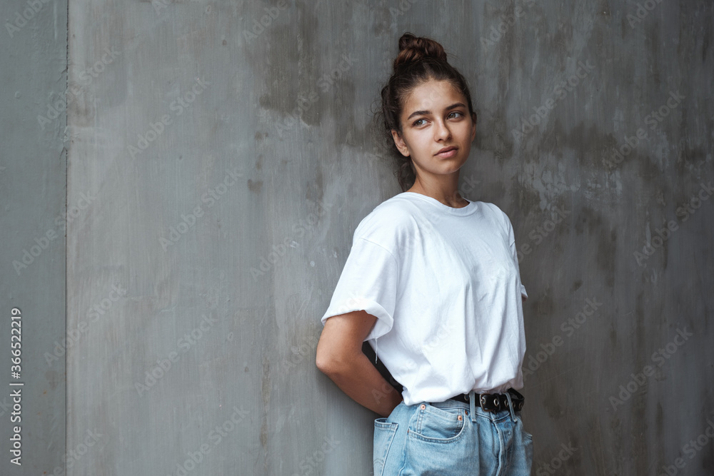 A sad pensive girl stands at the wall. Stylish young woman in blue jeans and a white T-shirt stands against the background of a gray concrete wall. A sad pensive girl stands at the wall. Stylish young woman in blue jeans and a white T-shirt stands against the background of a gray concrete wall.