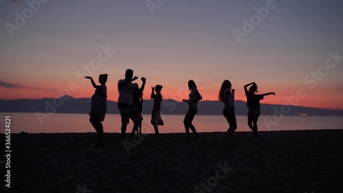 Silhouettes of cheerful dancing people on beautiful sea sunset background. Friends dancing and jumping, beach party and disco music. Fresh summer beverages for relax and holidays enjoyment photo
