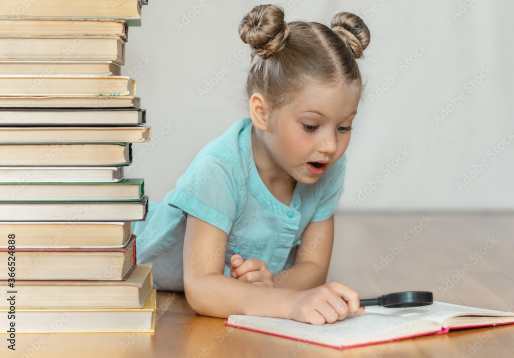 A little blonde schoolgirl looks at book through a magnifying glass in surprise, tearing her eyes off. A pile of books on blurred light background. Back to school. Education. Free time for the child. A little blonde schoolgirl looks at book through a magnifying glass in surprise, tearing her eyes off. A pile of books on blurred light background. Back to school. Education. Free time for the child.