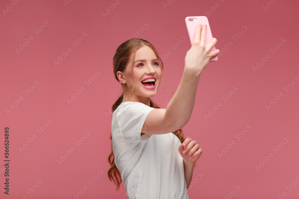 Funny young redhead woman making selfie. Smiling girl wearing white t-shirt holding pink smartphone, making faces on camera, posing for selfie isolated on pink background. Funny young redhead woman making selfie. Smiling girl wearing white t-shirt holding pink smartphone, making faces on camera, posing for selfie isolated on pink background.