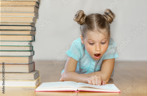 A little blonde schoolgirl reads and looks at the book in surprise, wide-eyed. A stack of books on a blurred light background. Back to school. Education. Free time for the child. Love of knowledge