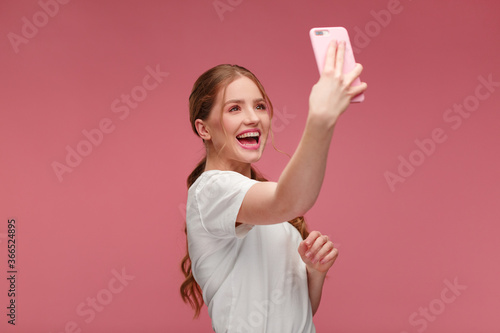 Funny young redhead woman making selfie. Smiling girl wearing white t-shirt holding pink smartphone  making faces on camera  posing for selfie isolated on pink background.