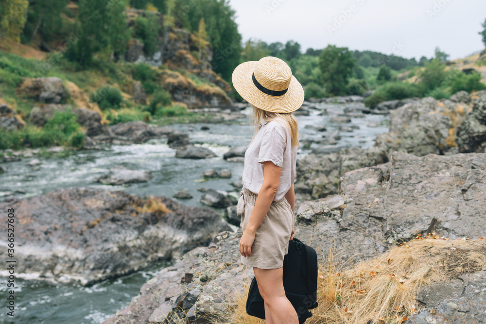 Young woman traveler in straw hat with backpack sits and looks at the mountain river, local travel concept