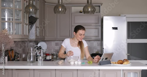 young woman cooking dough for pancakes or cookies and watching online recipe on a tablet in the kitchen.