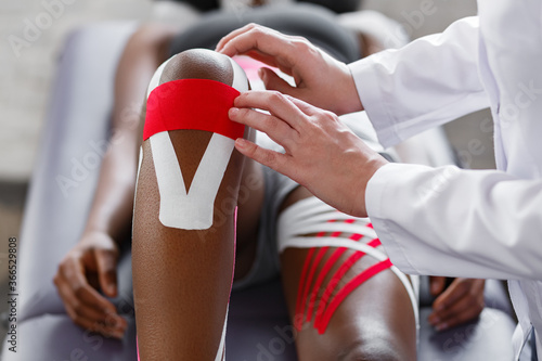 Physiotherapist applying kinesiology tape to patient knee.Therapist treating young female African American athlete. Kinesiology taping. Post traumatic rehabilitation,sport physical therapy.