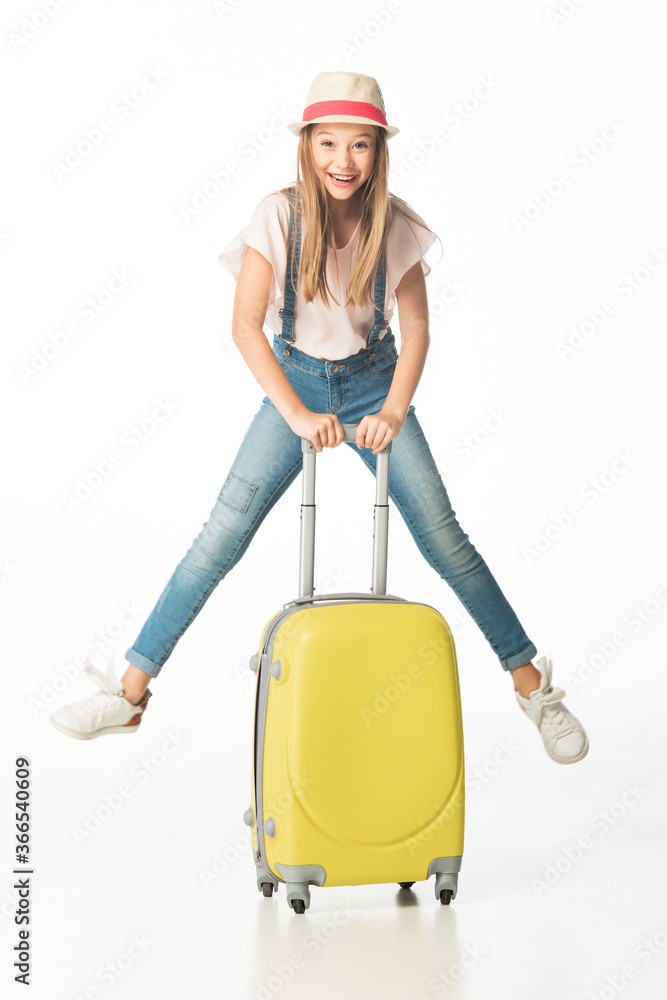 happy girl in hat jumping near yellow travel bag isolated on white happy girl in hat jumping near yellow travel bag isolated on white