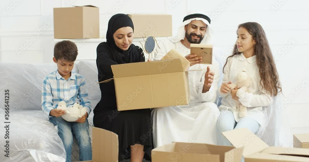 Cheerful Arabs parents sitting on sofa in living room with small kids and unpacking carton boxes. Muslims family with children moving in new house. Repairment. Adults and teens changing accomodation. Cheerful Arabs parents sitting on sofa in living room with small kids and unpacking carton boxes. Muslims family with children moving in new house. Repairment. Adults and teens changing accomodation.