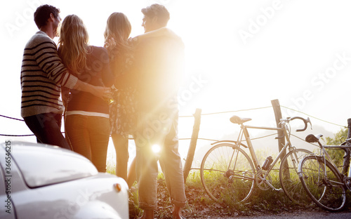 Four people standing by the side of a road, parked car and bicycles leaning against a fence, sunlight. photo