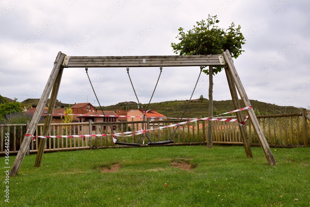 Fototapeta premium Swings in a playground in Spain closed by the Sars-Cov-2 pandemic (covid19). Its use is prohibited by placing plastic tape. In the background houses from the village of Clavijo.