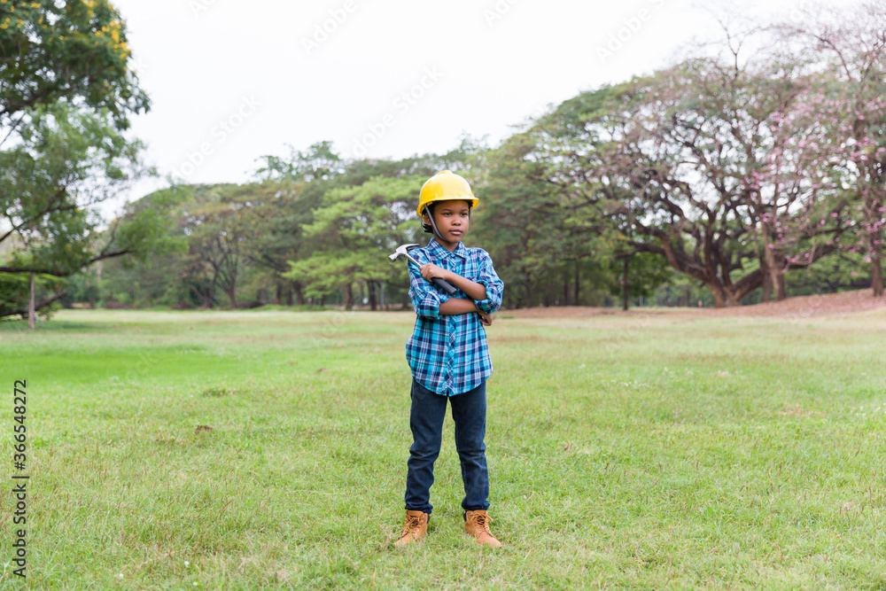 Portrait of African American young boy wearing engineer clothes and holding steel hammer, wearing yellow safety helmet standing in the garden while field trip or summer camp.