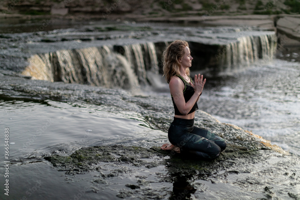 Blonde young woman in sportswear practicing breathing yoga outdoors in harmony with nature. Fitness girl meditating with closed eyes on the edge of waterfall with hands clasped in namaste mudra sign.