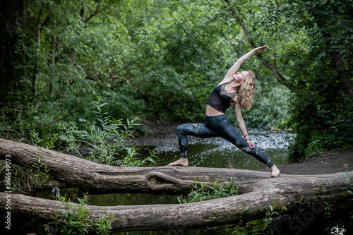 Blonde woman in sportswear practicing breathing yoga on a log over the forest river. Fitness girl doing a stretching exercise outdoor in harmony with nature.