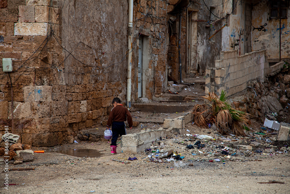 A scene from the back street slums of Tartus, Syria. It is a rundown dirty neighborhood with ruble and trash on the abandoned streets and ruined houses. A boy is seen carrying food in a plastic cup.