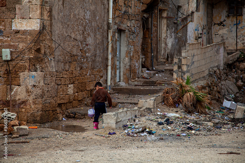A scene from the back street slums of Tartus, Syria. It is a rundown dirty neighborhood with ruble and trash on the abandoned streets and ruined houses. A boy is seen carrying food in a plastic cup. photo