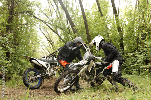 Strong men in helmets picking up motorcycle together in forest while practicing off-road skills