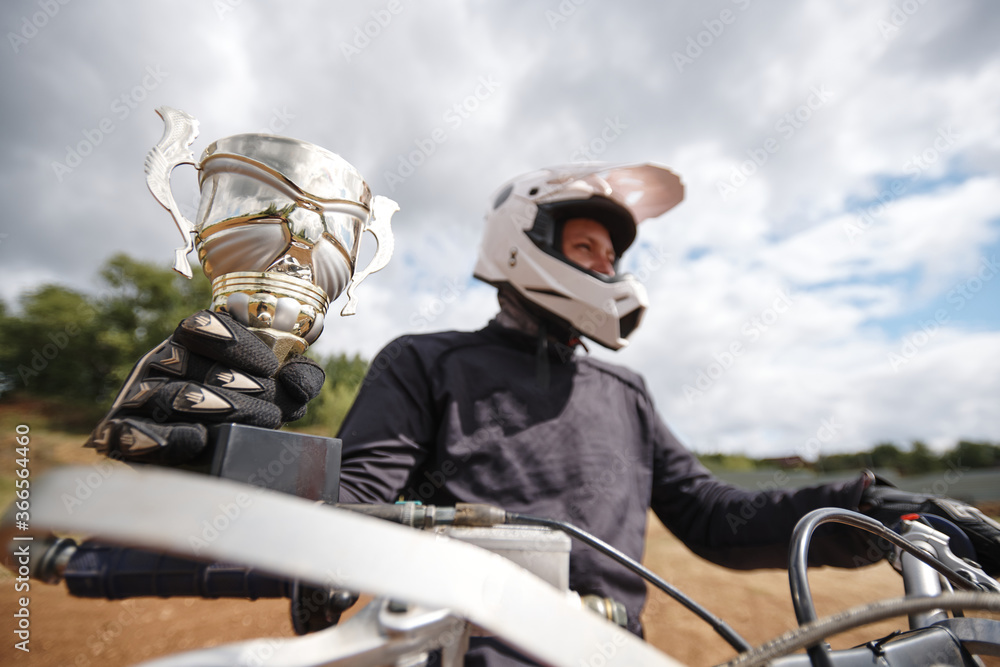 Close-up of successful motorcyclist in helmet winning motorcycle championship riding motorbike and holding cup
