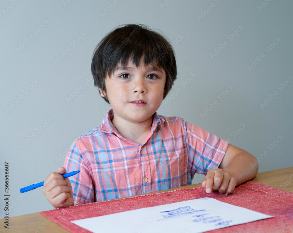 Portrait of school kid boy siting on table doing homework, Happy Child holding blue colour paint,A boy drawing blue colour on white paper at the table,Elementary school and education concept Portrait of school kid boy siting on table doing homework, Happy Child holding blue colour paint,A boy drawing blue colour on white paper at the table,Elementary school and education concept