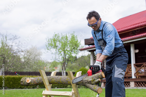 Male worker in overalls saws a tree on sawhorses in courtyard with a chainsaw. Cutting firewood in forest with a professional chainsaw. Hard work with a saw. Sawdust fly to sides