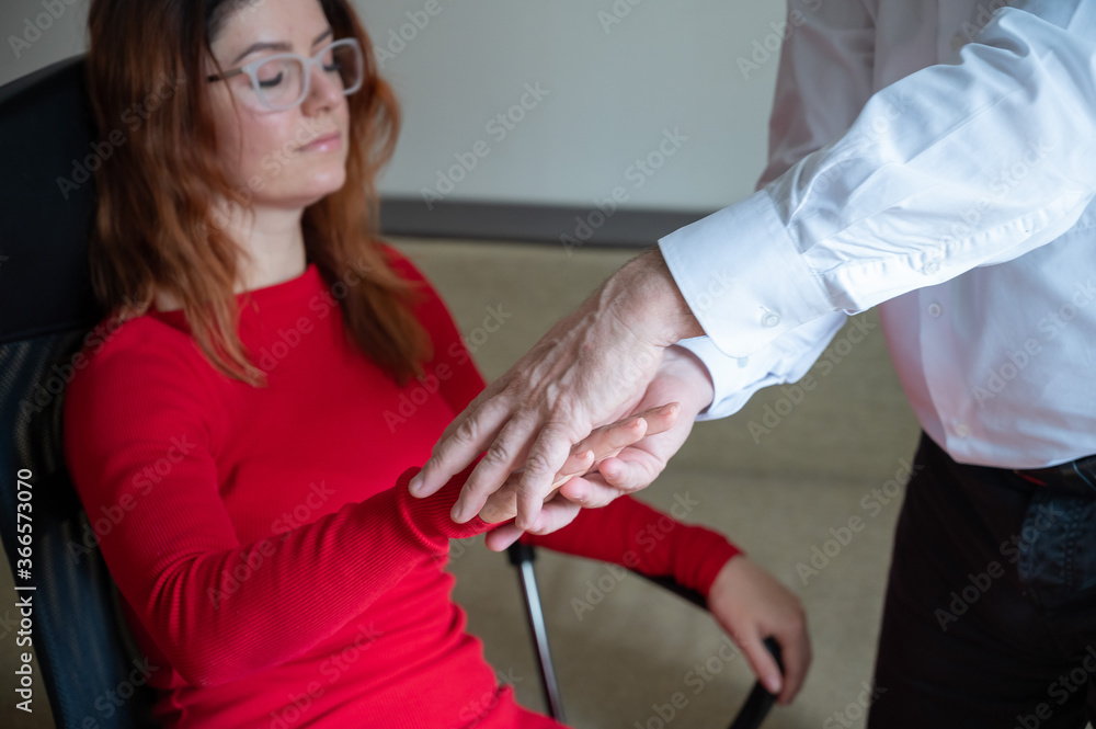 A red-haired woman sits in a chair with her hands closed and hands raised during gopnosis. Mature male hypnotherapist in session with a female patient. Alternative medicine for mental health. Cropped.