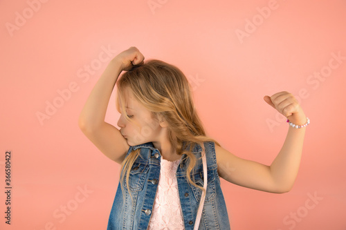 Portrait of funny cheerful little caucasian schoolgirl kissing biceps and showing strength, pink background, copy space, kids healthy lifestyle concept