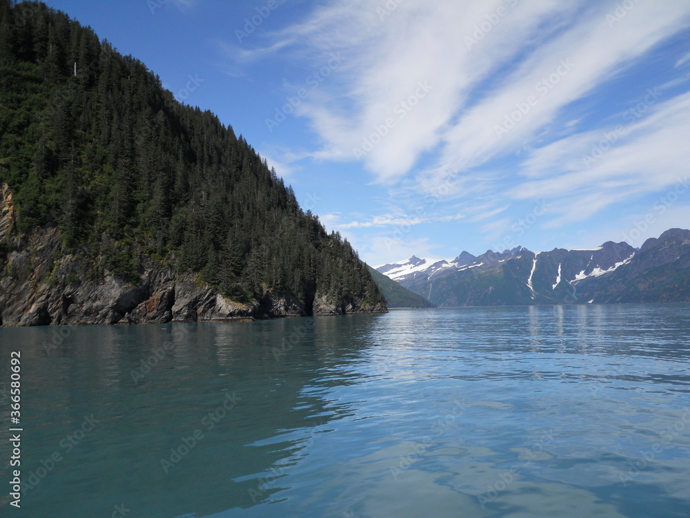 Alaskan Bay Coastline Island View with Reflection in Ocean Water, Conifer Covered Rocky Mountains, and White Clouds in a Blue Sky