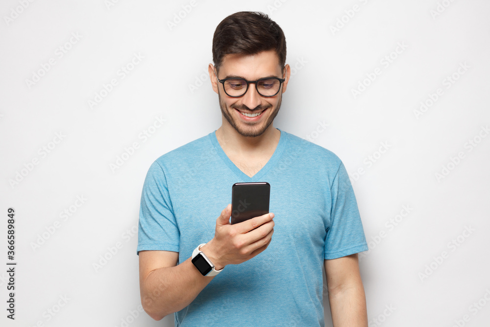 Young handsome man in blue t shirt holding smartphone and looking at phone screen with smile while standing, isolated on gray background Young handsome man in blue t shirt holding smartphone and looking at phone screen with smile while standing, isolated on gray background