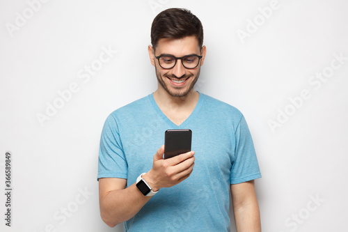 Young handsome man in blue t shirt holding smartphone and looking at phone screen with smile while standing, isolated on gray background © Damir Khabirov