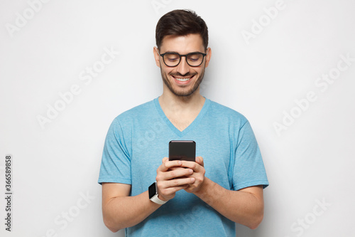 Portrait of young handsome guy wearing blue-shirt and glasses looking at phone screen with smile, isolated on gray background © Damir Khabirov