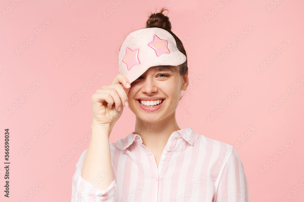 Young happy smiling girl wearing eye mask for sleeping, keeping it on forehead and winking with one eye, isolated on pink background Young happy smiling girl wearing eye mask for sleeping, keeping it on forehead and winking with one eye, isolated on pink background