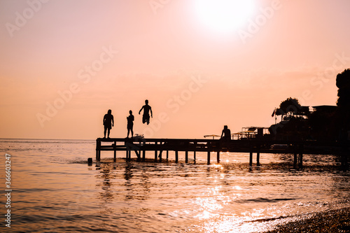 Young people as silhouette jumping over the sea from a wooden pier and having fun at sunset