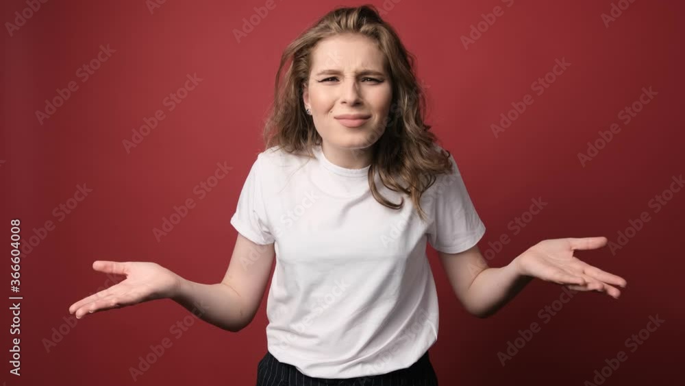 What wrong?! Portrait of confused beautiful girl with curly hair, spreads hands sideways, smirks face, feels doubt while makes choice, dressed in casual t shirt. Isolated on red background. What wrong?! Portrait of confused beautiful girl with curly hair, spreads hands sideways, smirks face, feels doubt while makes choice, dressed in casual t shirt. Isolated on red background.
